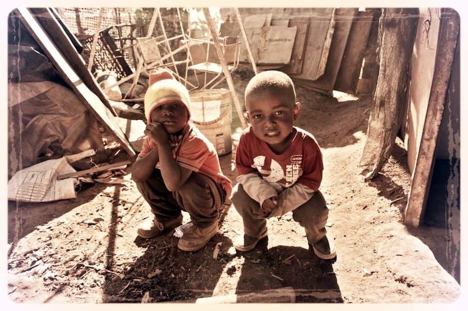 Two children crouch in a sunlit rustic wooden shelter, looking toward the camera.