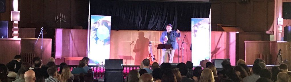 Speaker on a lit stage addressing an audience in a large auditorium with pink lighting