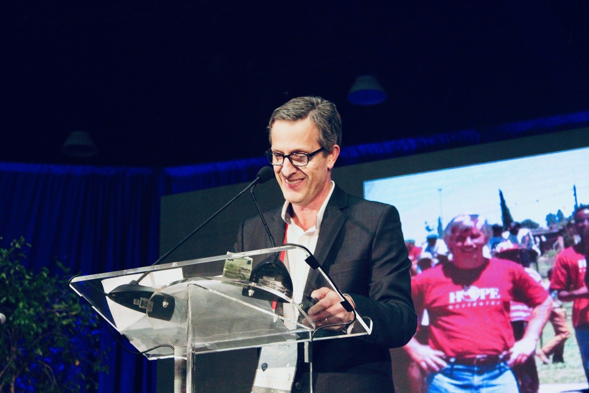 Man speaking at a clear podium on a stage, with a large screen showing people behind him.