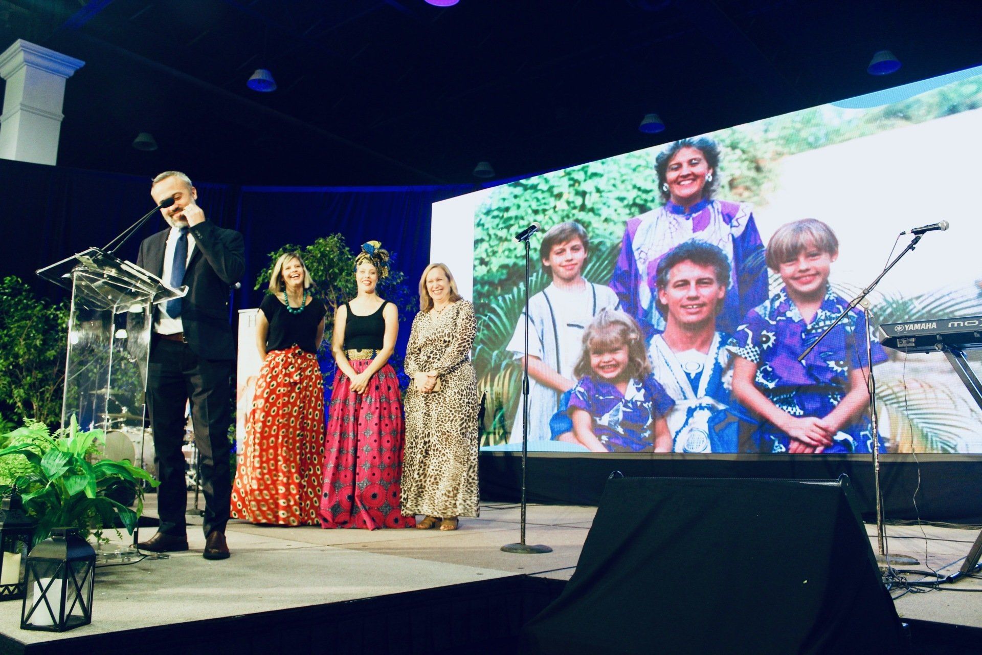 Award presentation on stage with four people speaking and a large family photo displayed on a screen