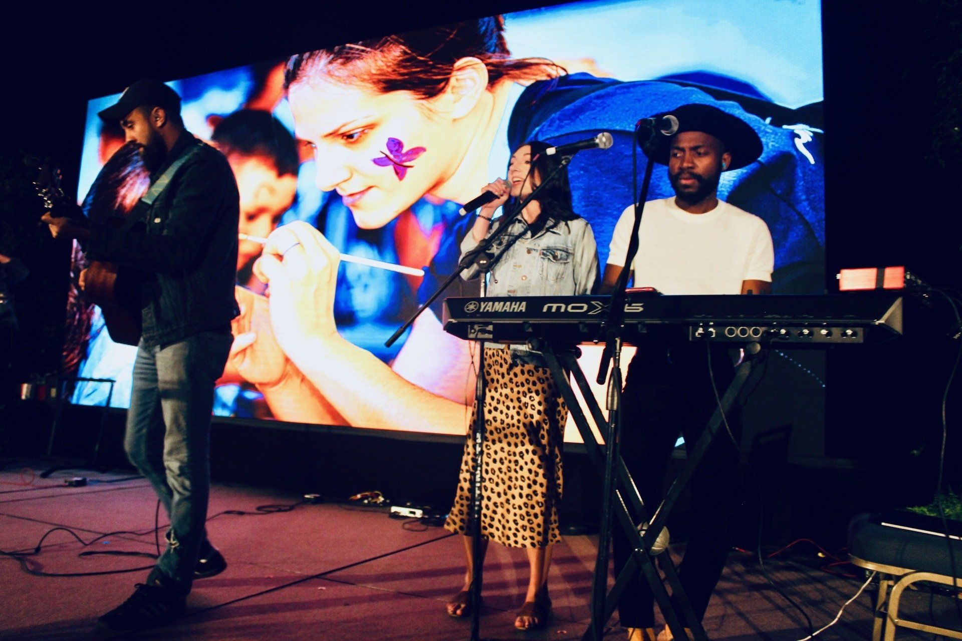 Three performers on stage, with a keyboardist, singer, and large colorful video screen behind them