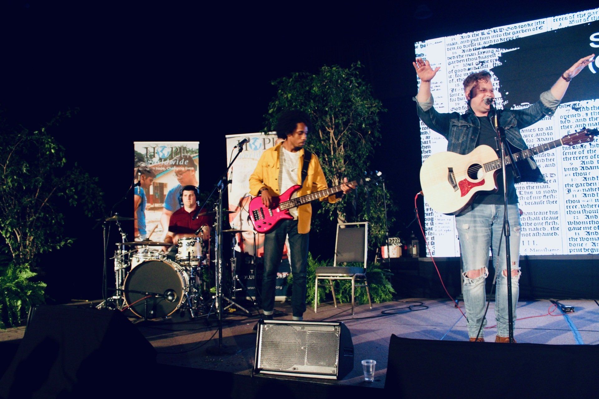 Band performs on an outdoor stage at night, with guitar, drums, and a bright LED backdrop.