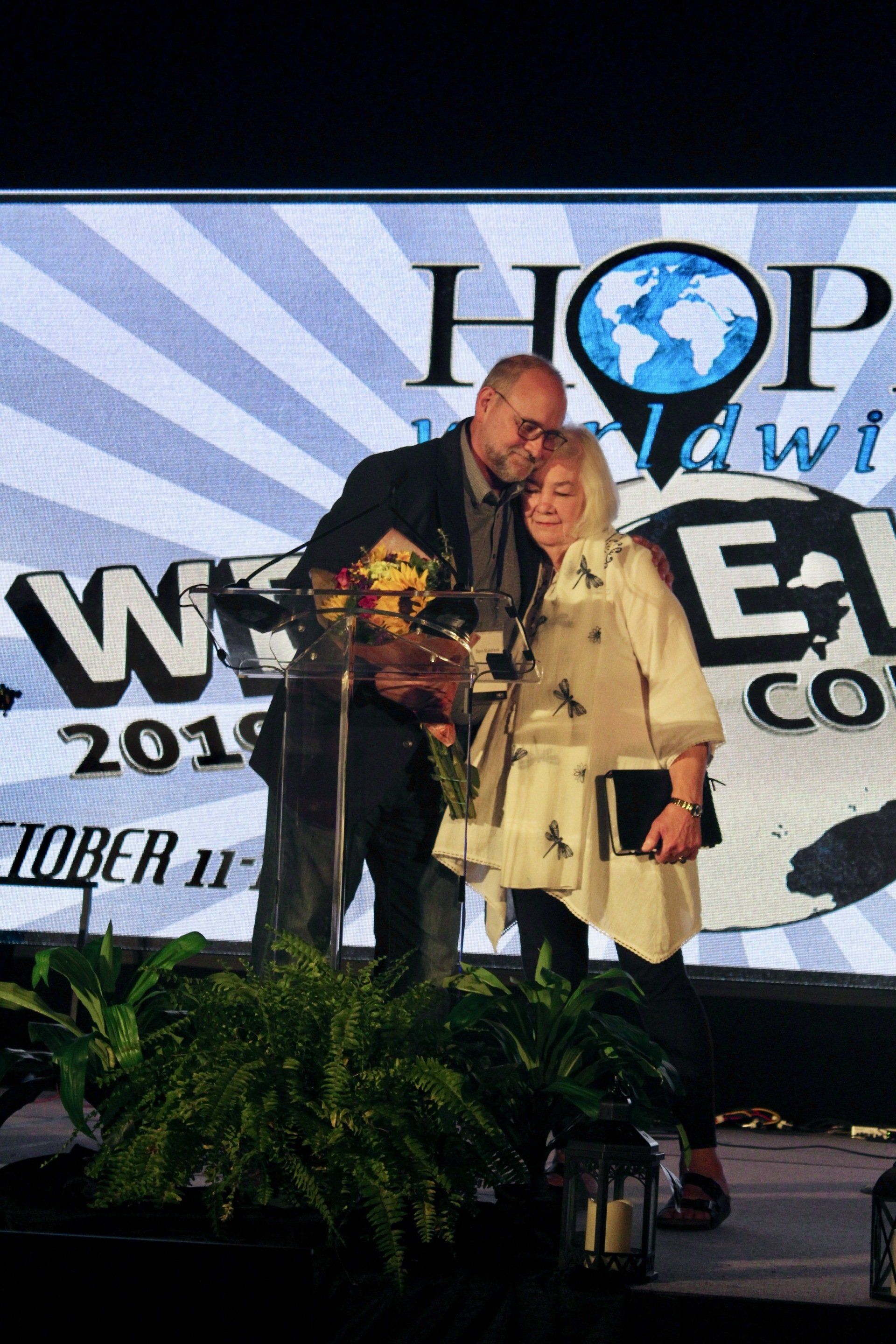 Two people on stage with a trophy, in front of a “HOP Hawaii” event backdrop and potted plants.