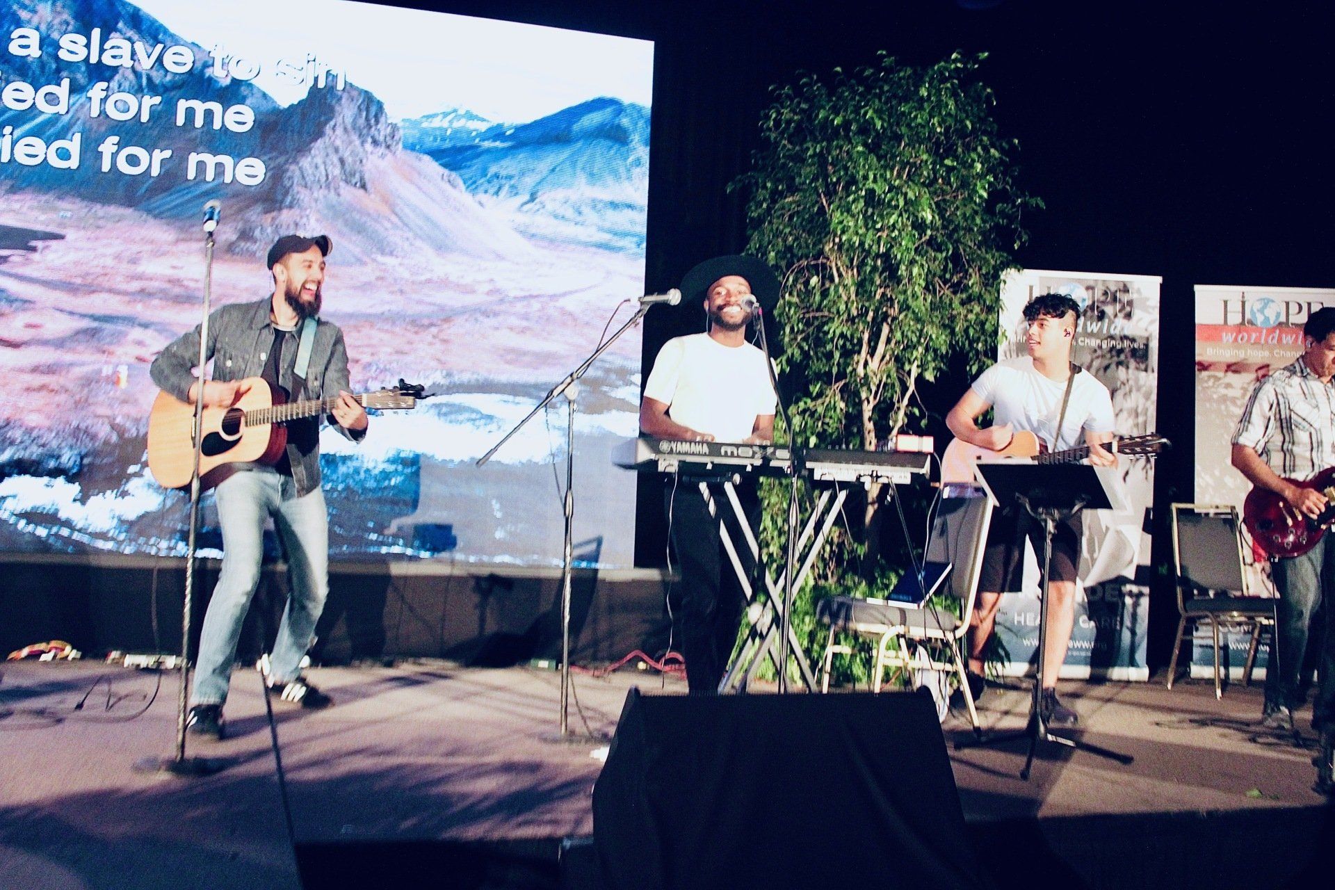 Three musicians perform on an outdoor stage, with guitars, keyboard, and a mountain backdrop.