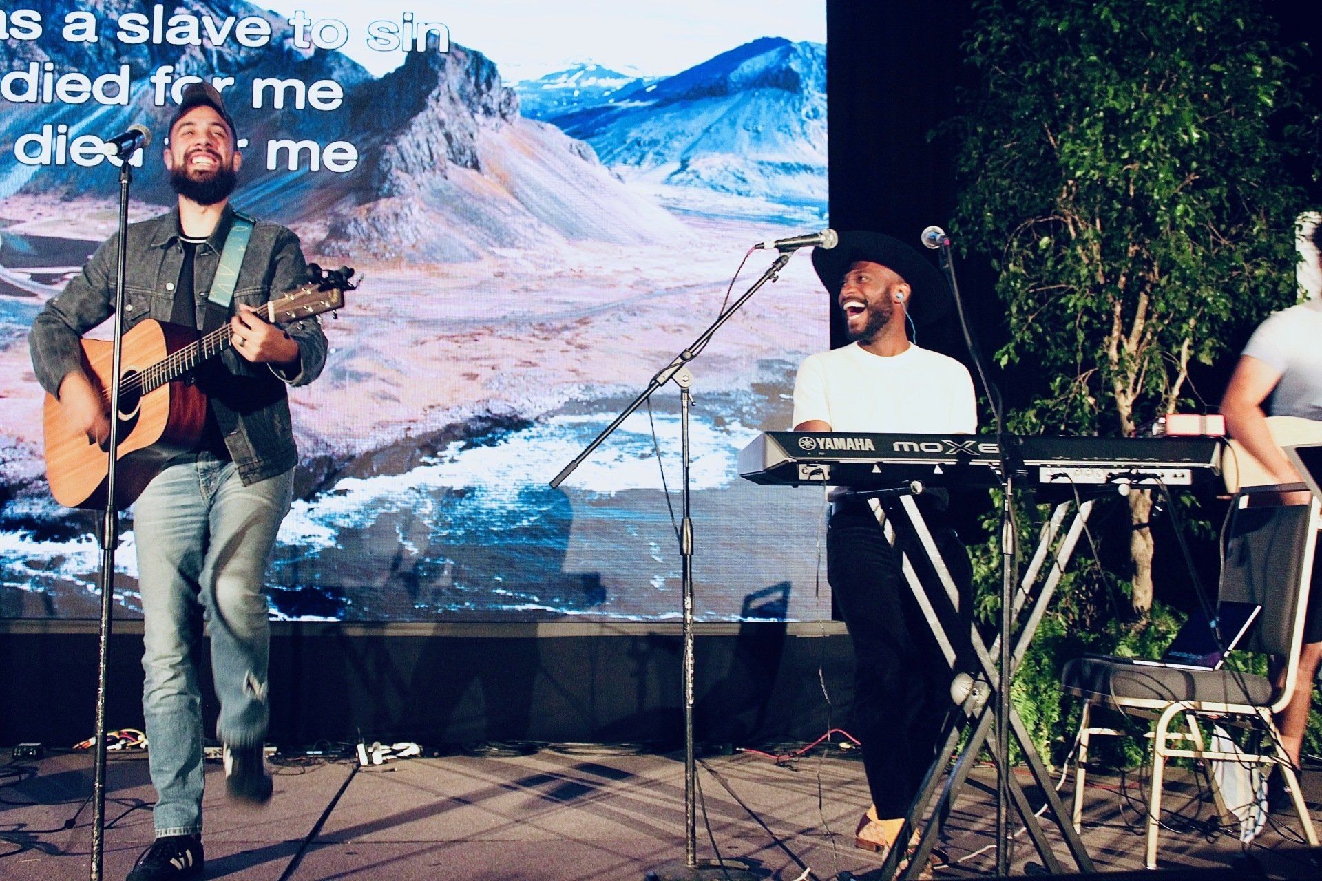 Musicians performing on an outdoor stage, with a guitarist, keyboard player, and mountain backdrop on screen