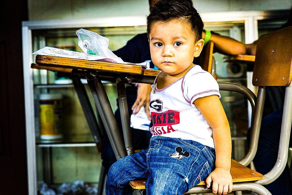 Child sitting on a chair at a table, holding the edge and looking at the camera.