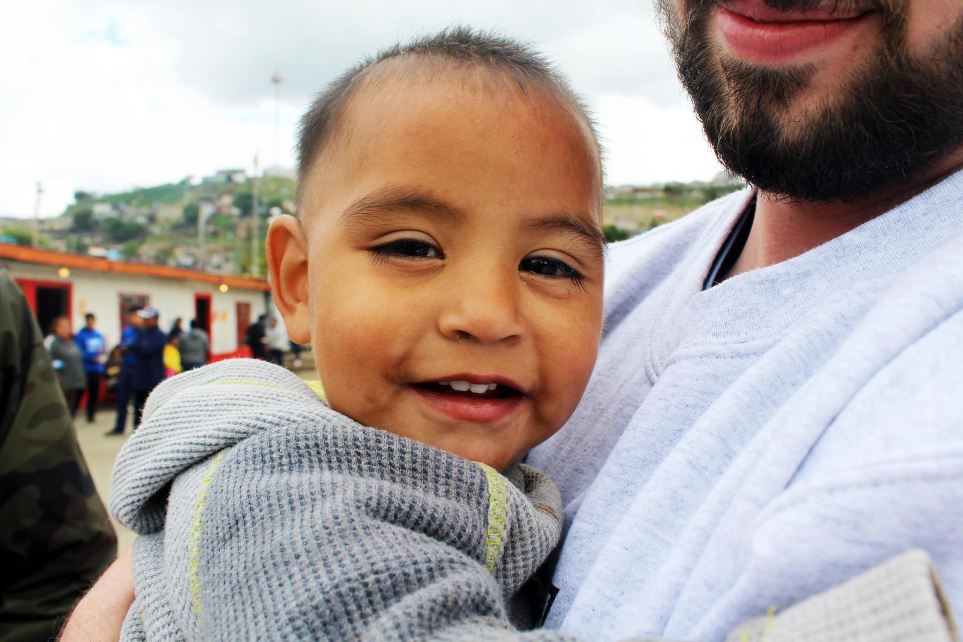 Smiling child in a gray sweater being held by an adult outdoors, with a blurred crowd in the background