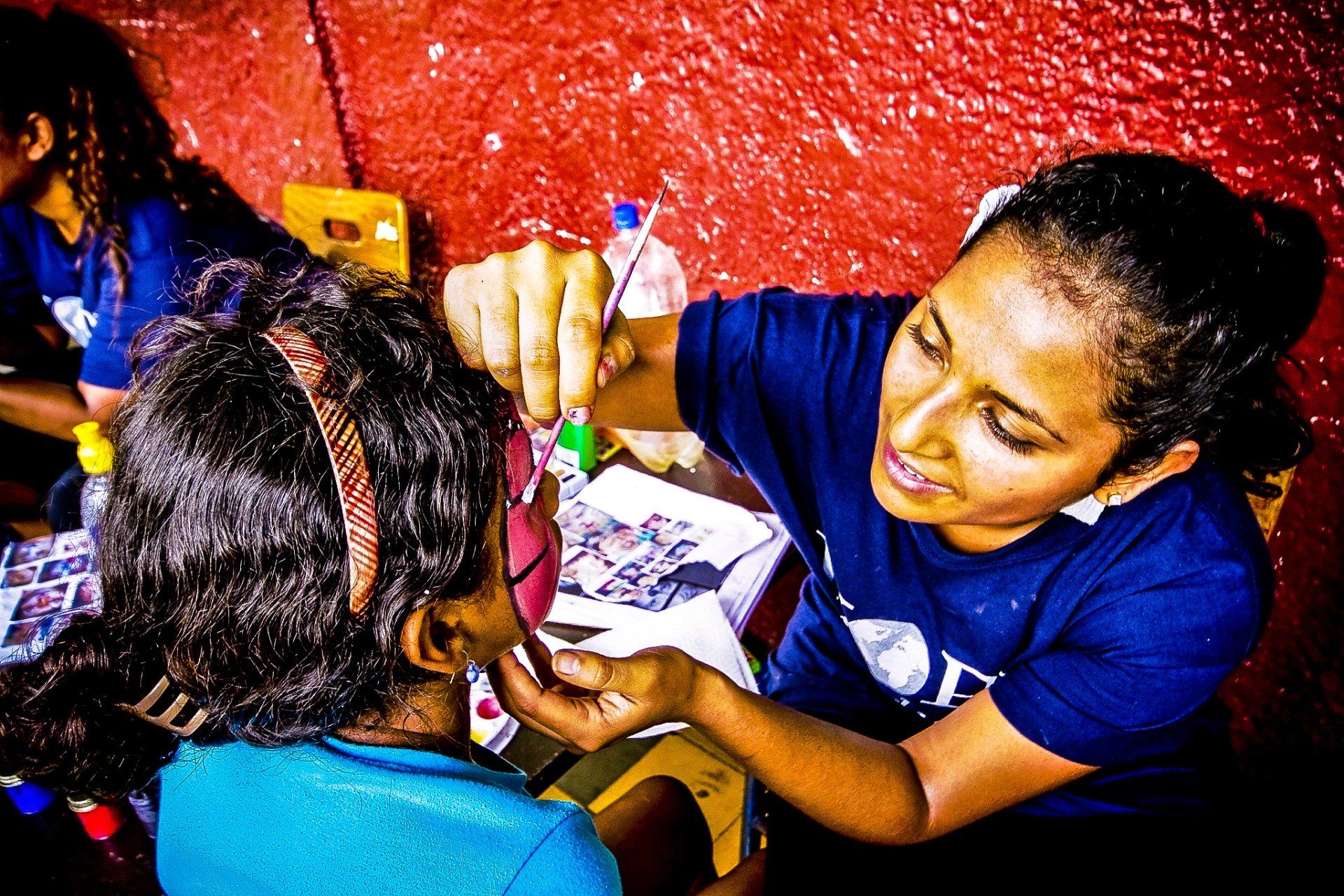 Woman painting another person’s face at a table, with art supplies and a red wall in the background