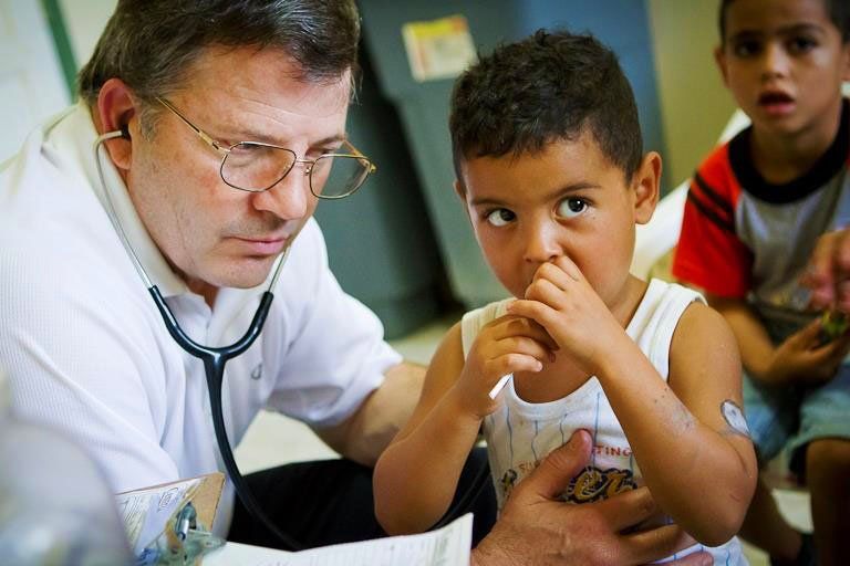 Doctor examining a child in a clinic while another child watches nearby