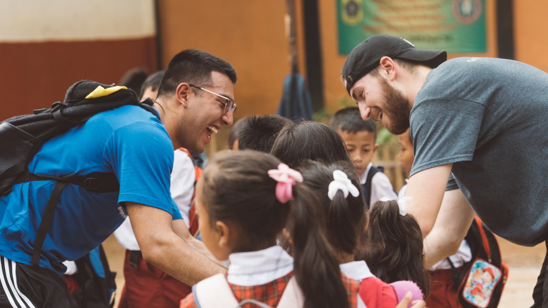 Two adults leaning in and talking with children in a classroom, backpacks visible.