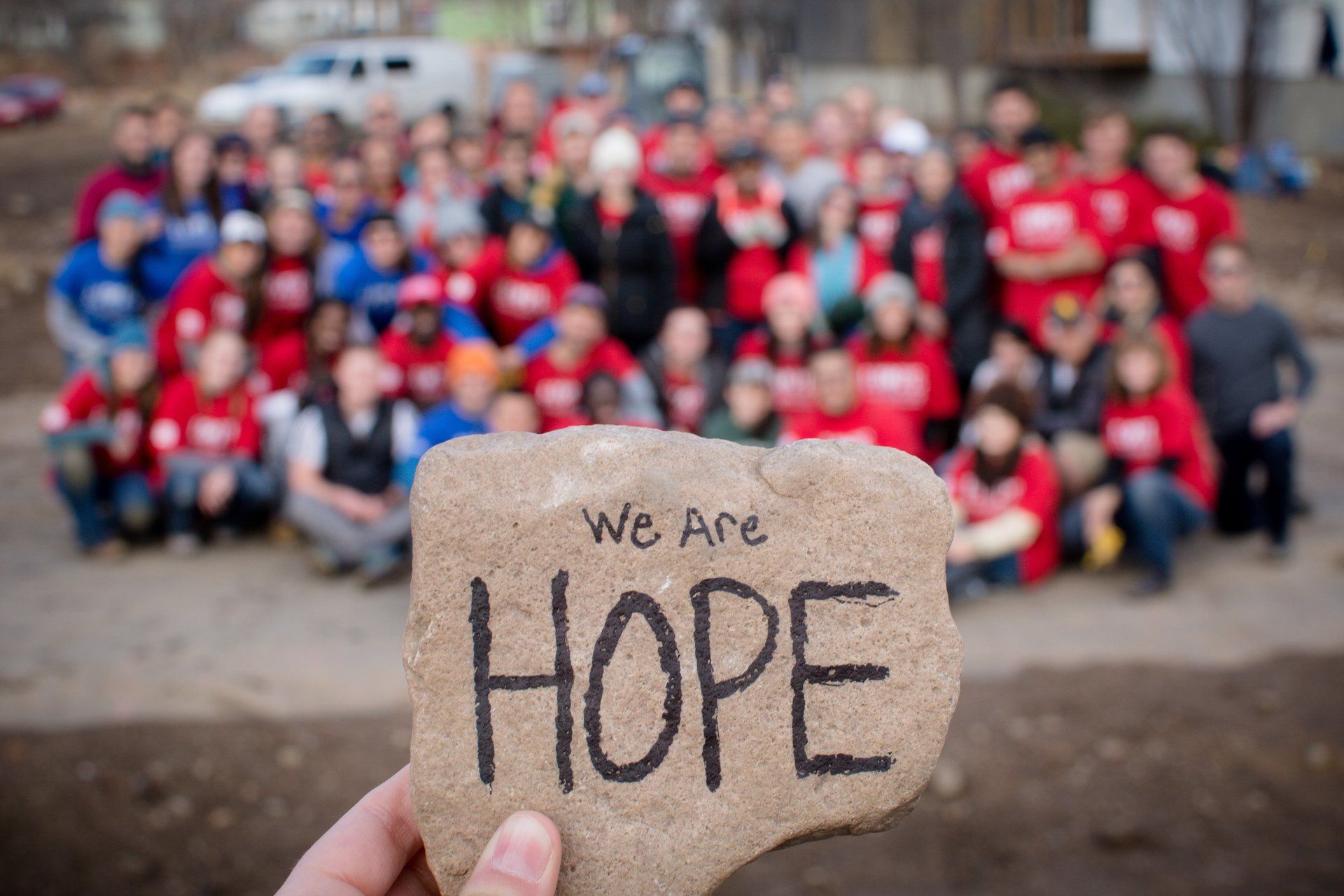 Hand holding a stone reading “We are HOPE” with a blurred crowd in red shirts behind it