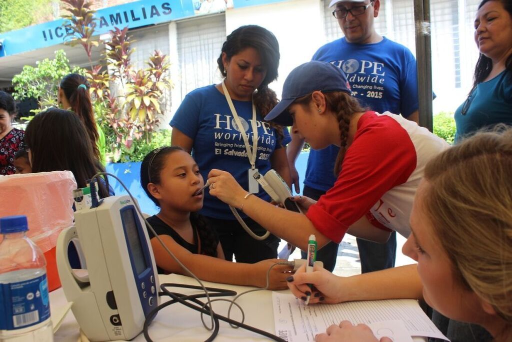 Health fair volunteers check a seated girl’s blood pressure at a table under a blue canopy.