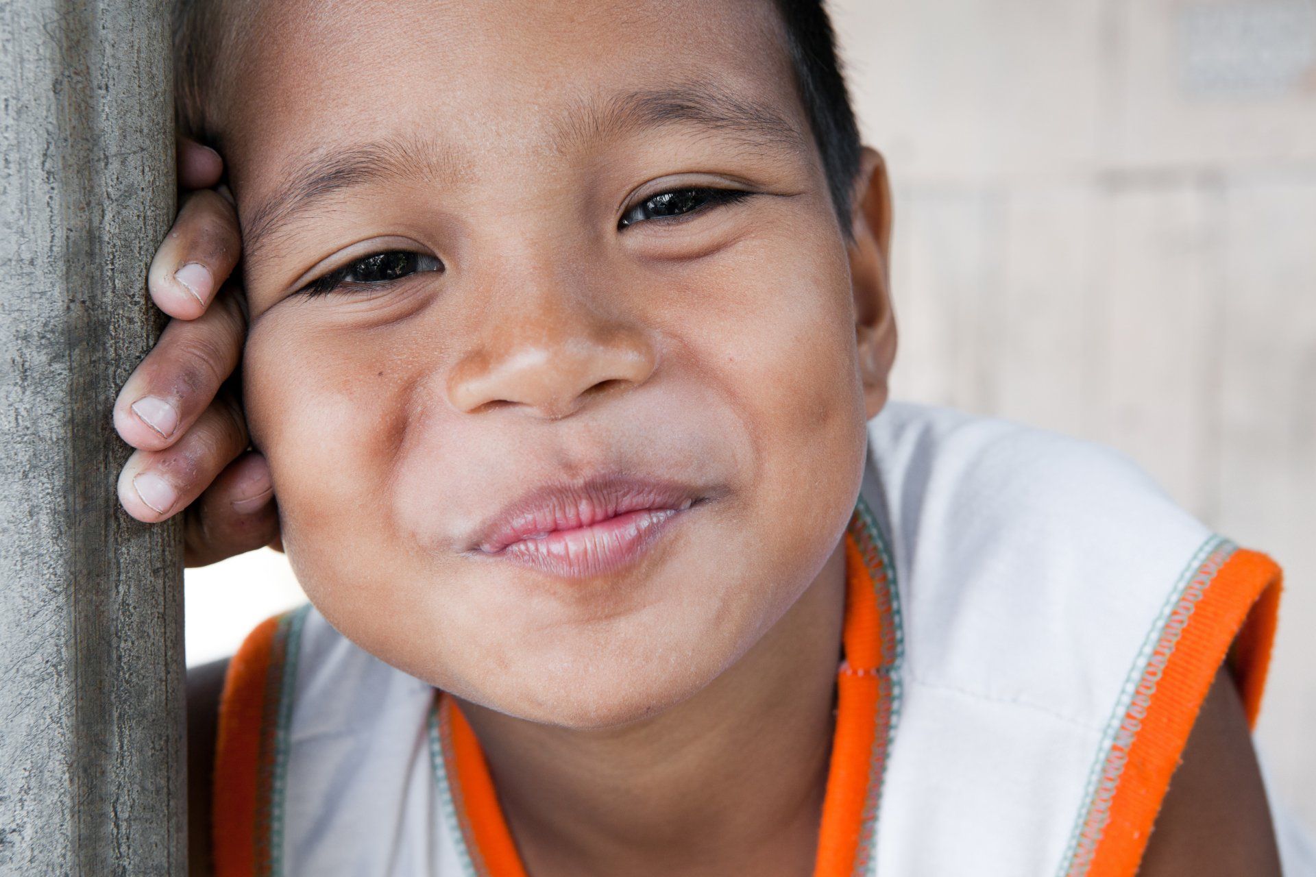 Smiling child leaning against a wooden post, wearing a white shirt with orange trim.