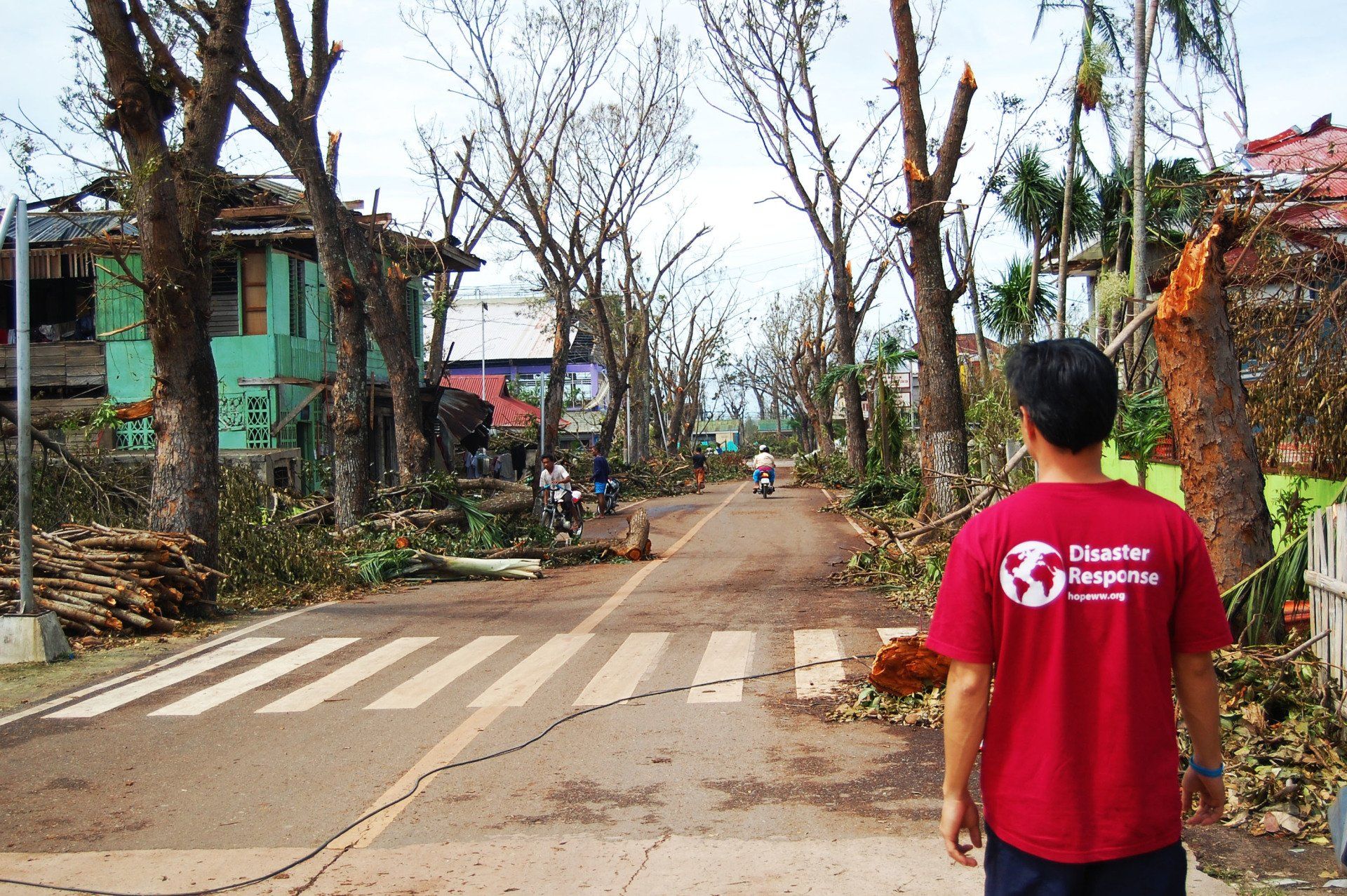 Person in red shirt stands on a debris-strewn street with damaged trees and houses after a storm