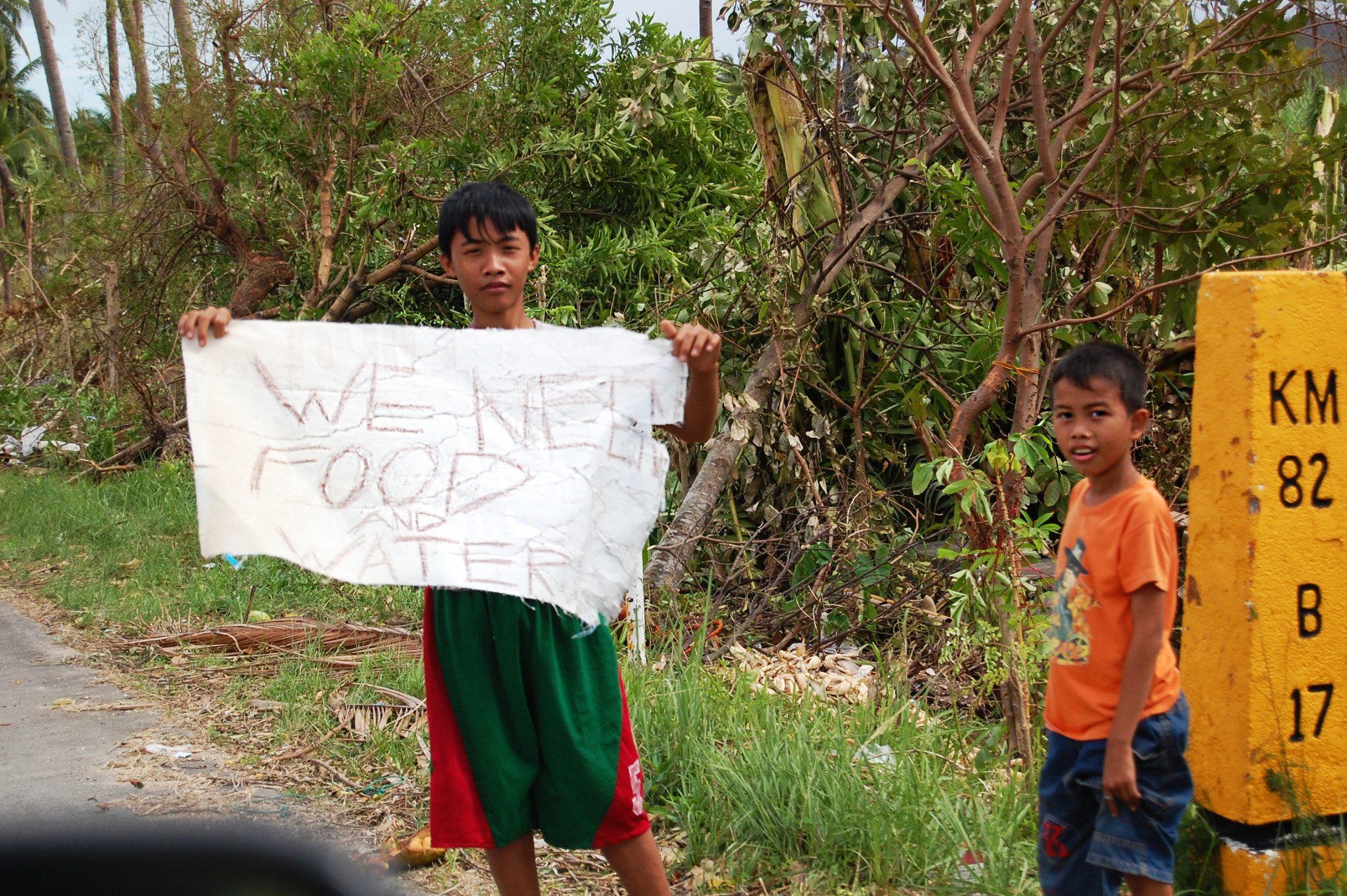 Two children stand by a roadside; one holds a handwritten protest sign, with a kilometer marker nearby.