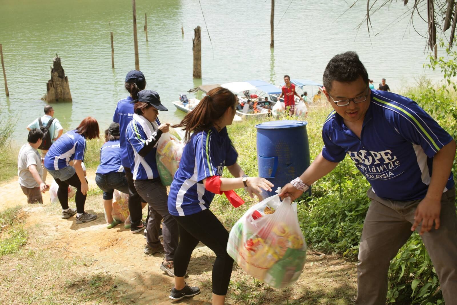 Volunteers in blue shirts cleaning trash along a lake shore, carrying bags of litter
