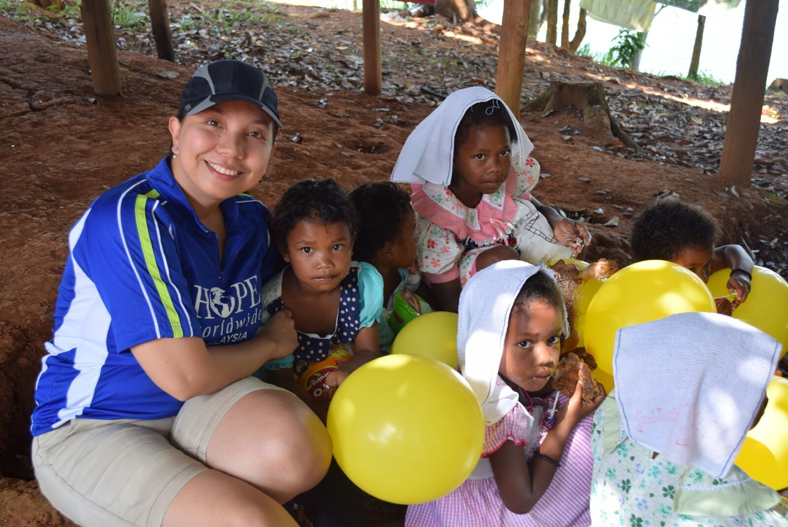 Smiling volunteer sits with children holding yellow balloons under a shaded shelter