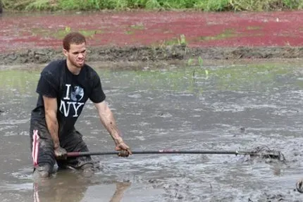 Man in a black shirt wades through muddy water while holding a long pole in a flooded area