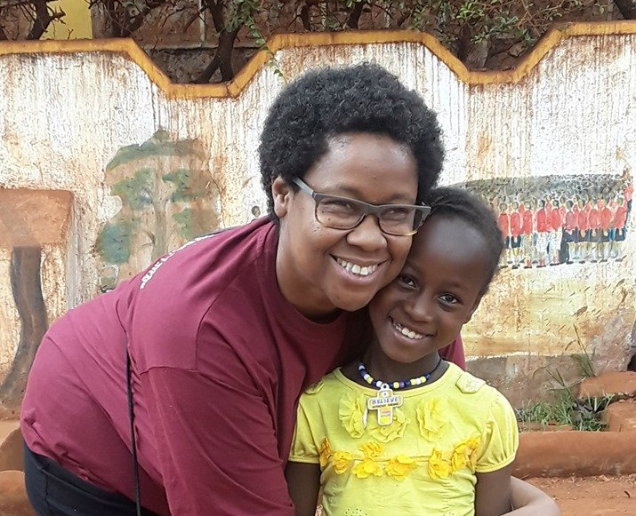 Smiling woman in maroon top hugging a child in a yellow shirt outdoors against a weathered wall
