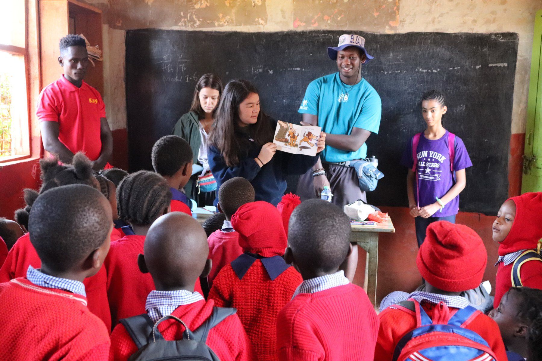Teacher and volunteers distribute supplies to students in a classroom with a chalkboard.