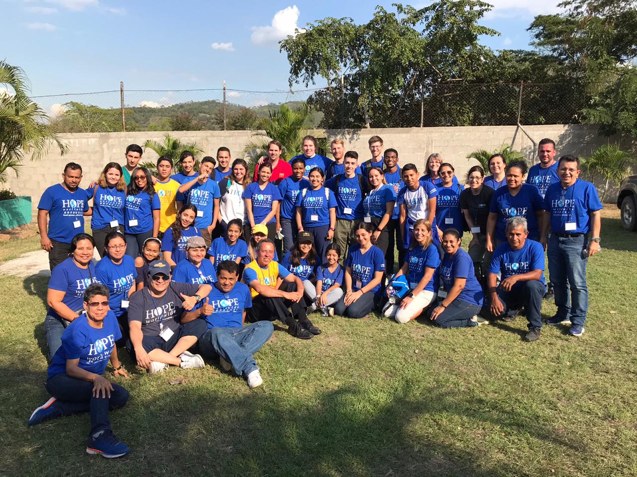 Large group posing outdoors on grass, many in blue shirts, with trees and hills in the background