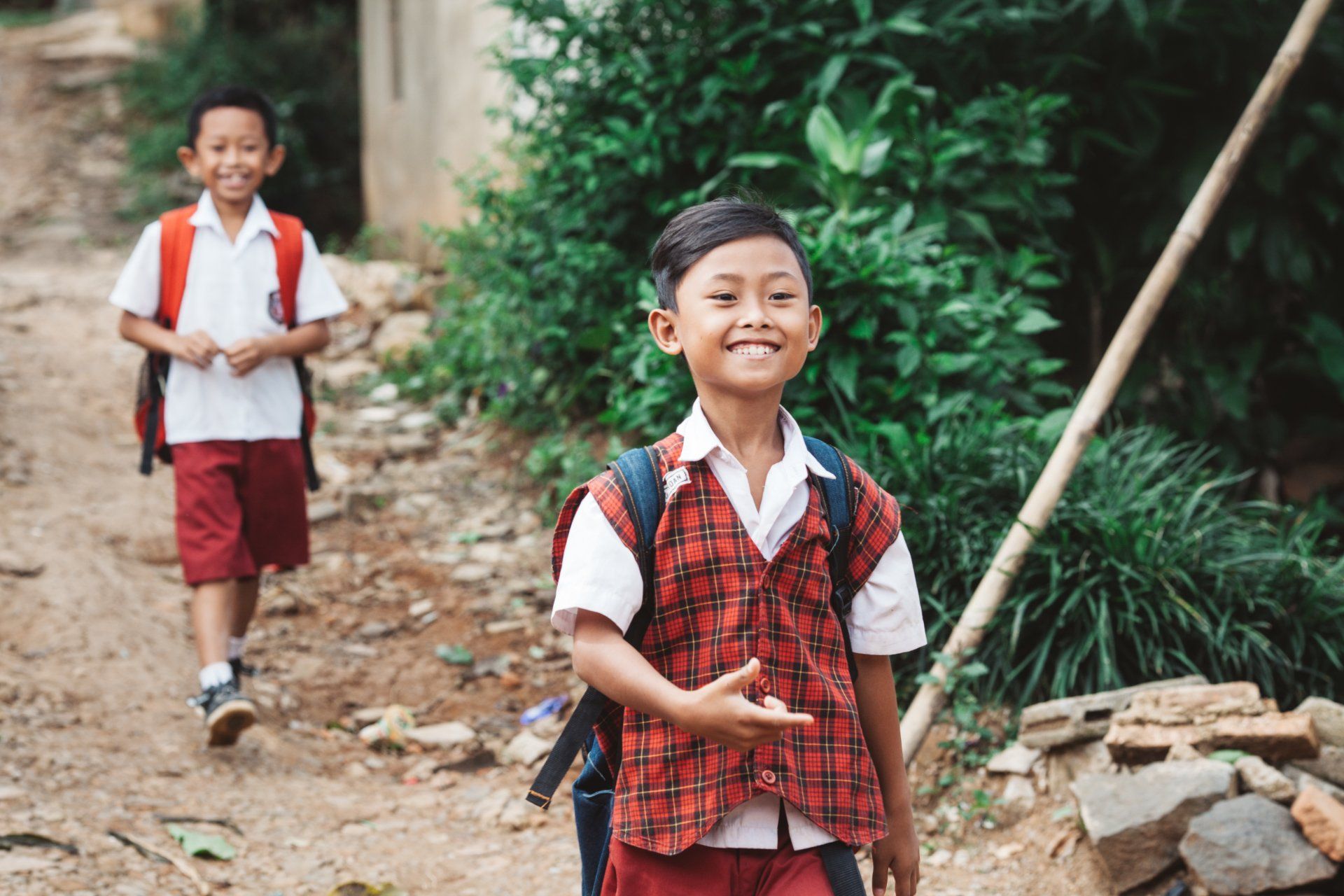 Two children in red school uniforms walk along a dirt path, one smiling at the camera.
