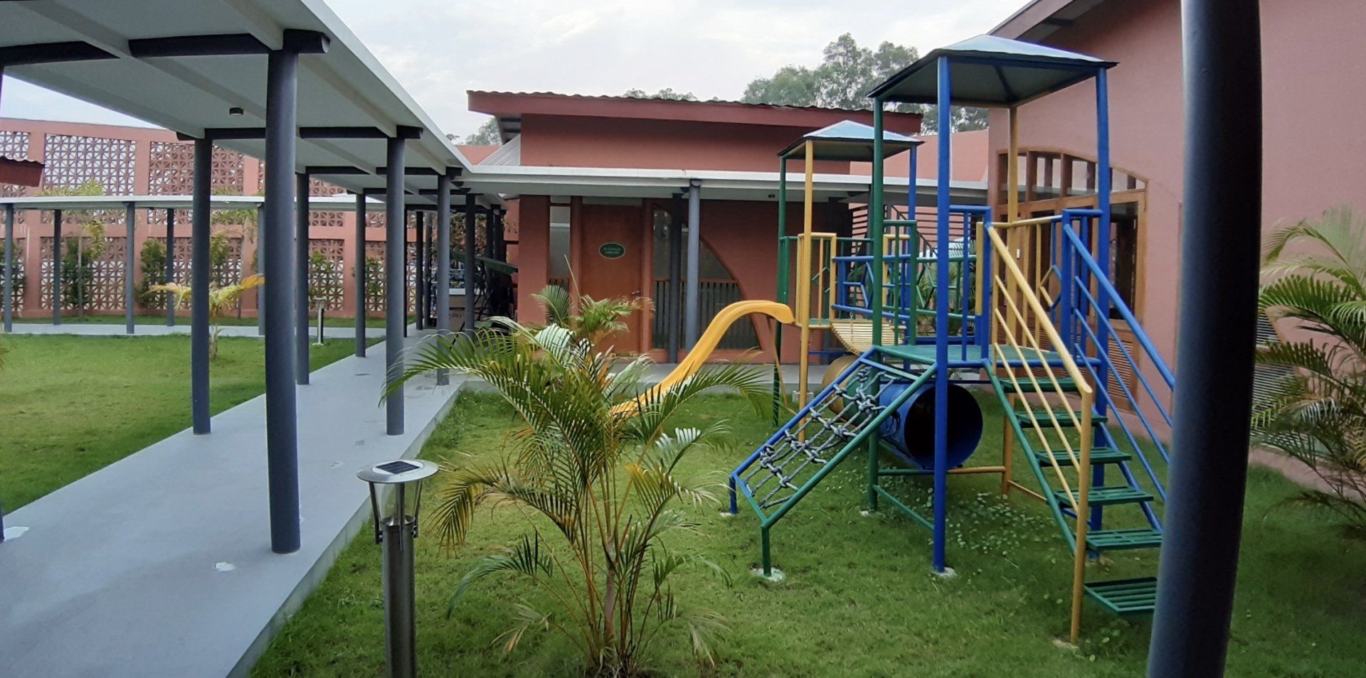 Covered walkway beside a small playground with blue climbing frames, a slide, and grassy courtyard