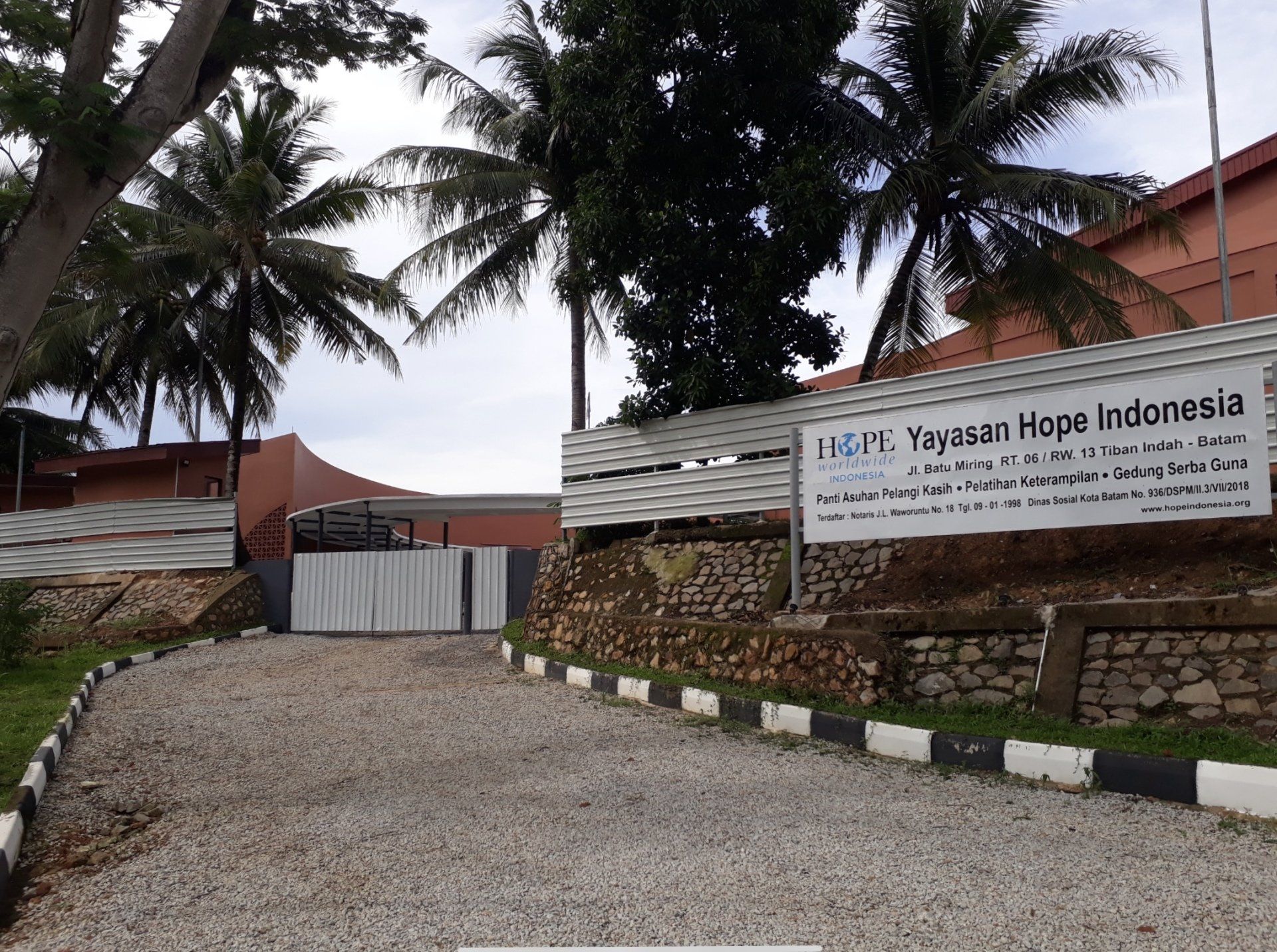 Entrance gate to Yayasan Hope Indonesia, with palm trees, gravel drive, and stone walls.