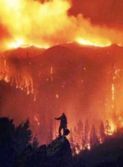 Silhouette of a person on a rocky outcrop against a blazing forest fire at night