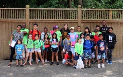 Large group of children and adults posing outdoors in front of a wooden fence, some holding bags and papers