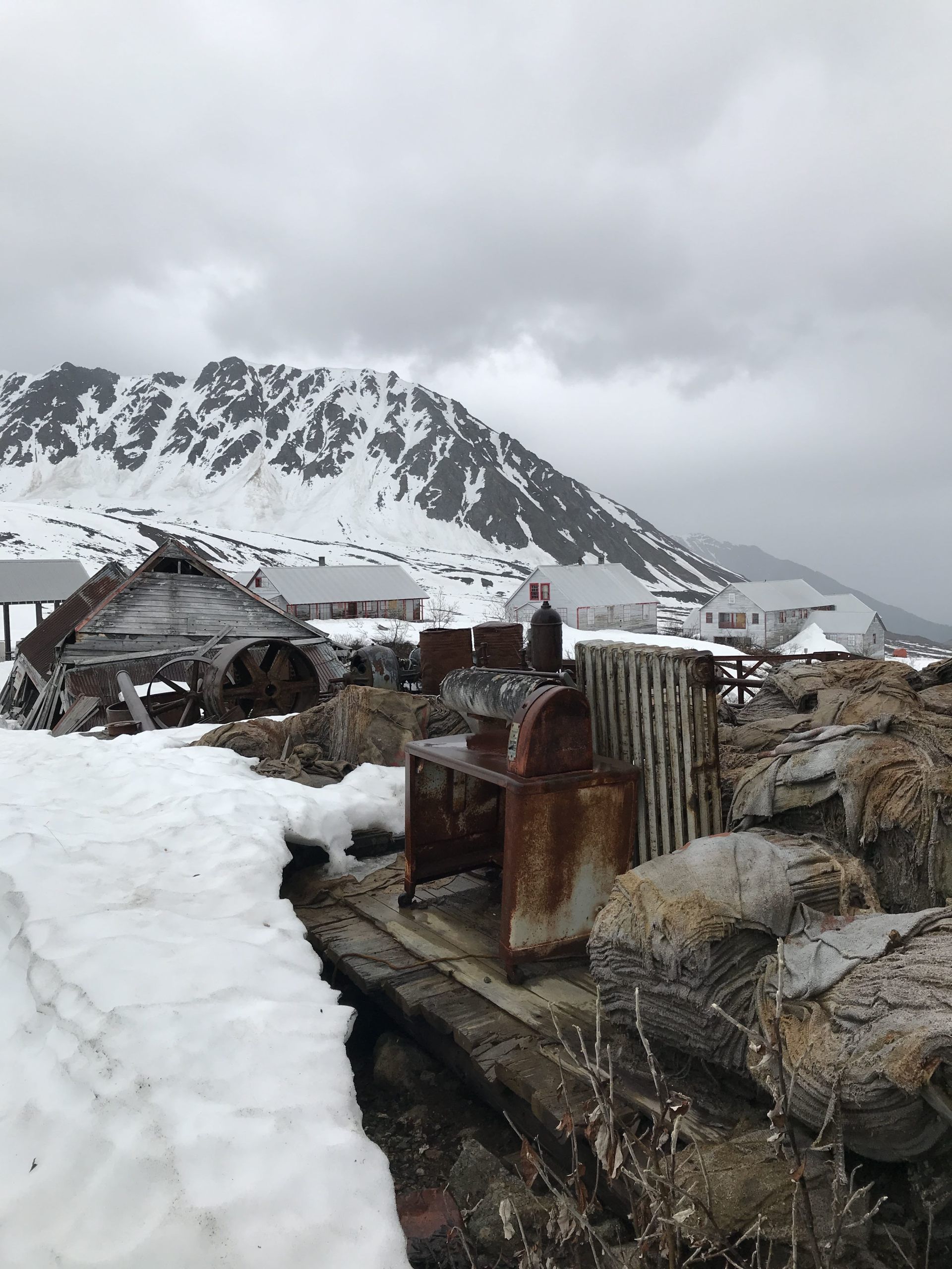 Snowy coastal settlement with rusted machinery in the foreground and mountains under gray clouds in the background