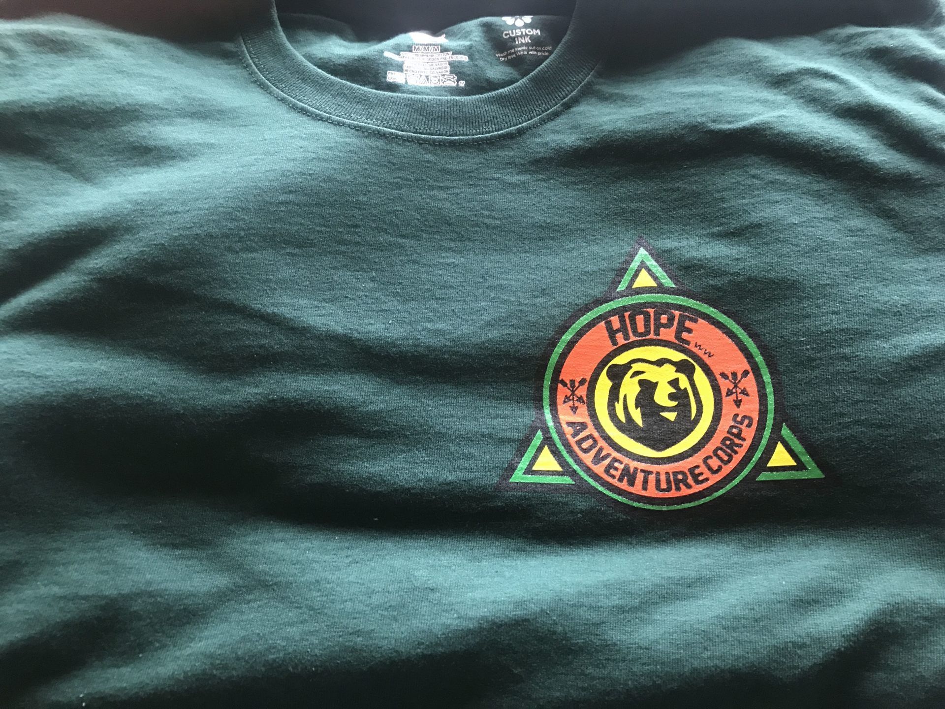 Dark green T-shirt with a colorful circular and triangular mountain-themed patch on the chest.