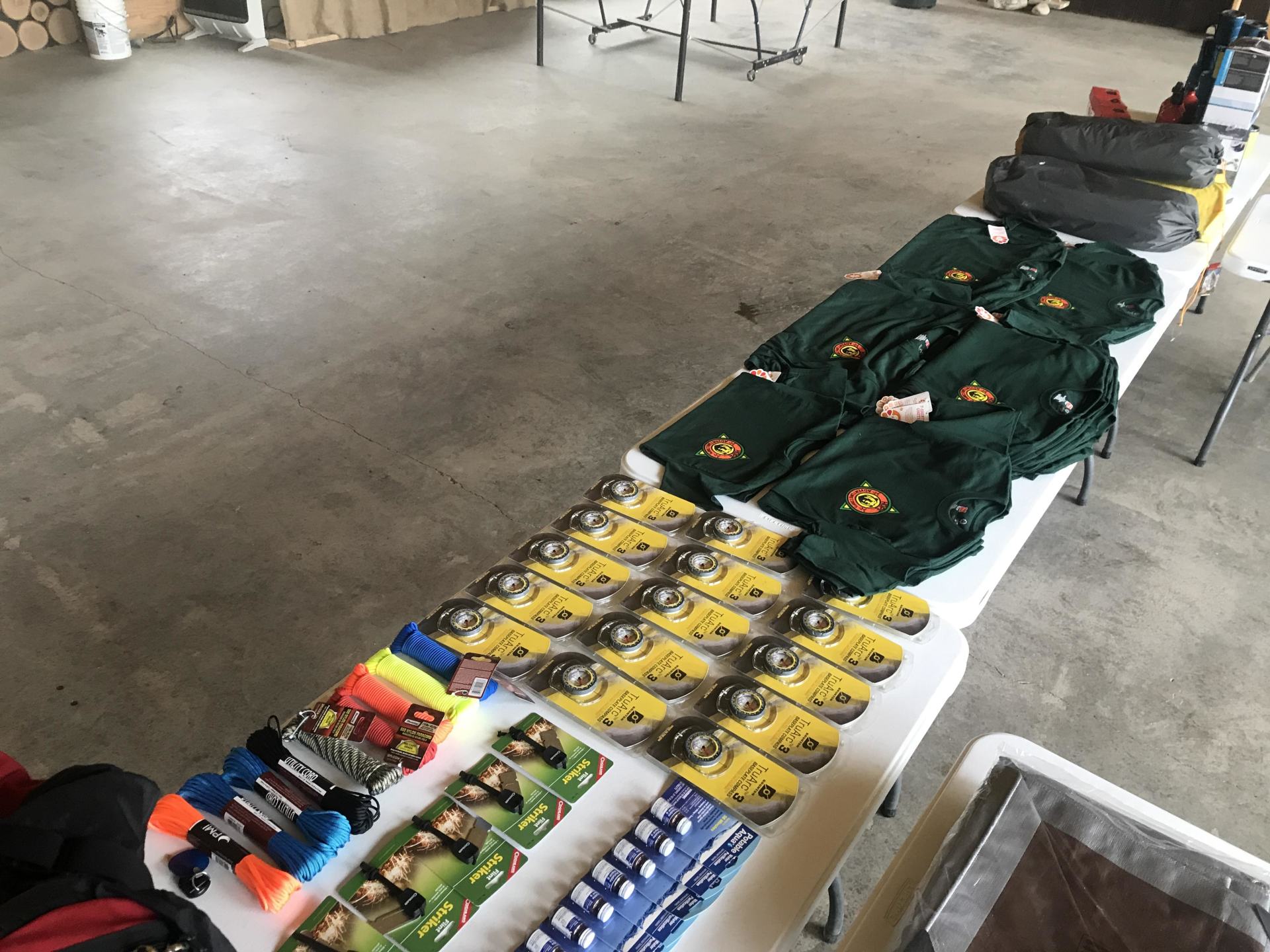 Table covered with rows of yellow packaged items, folded green clothing, and assorted colorful supplies in a garage.