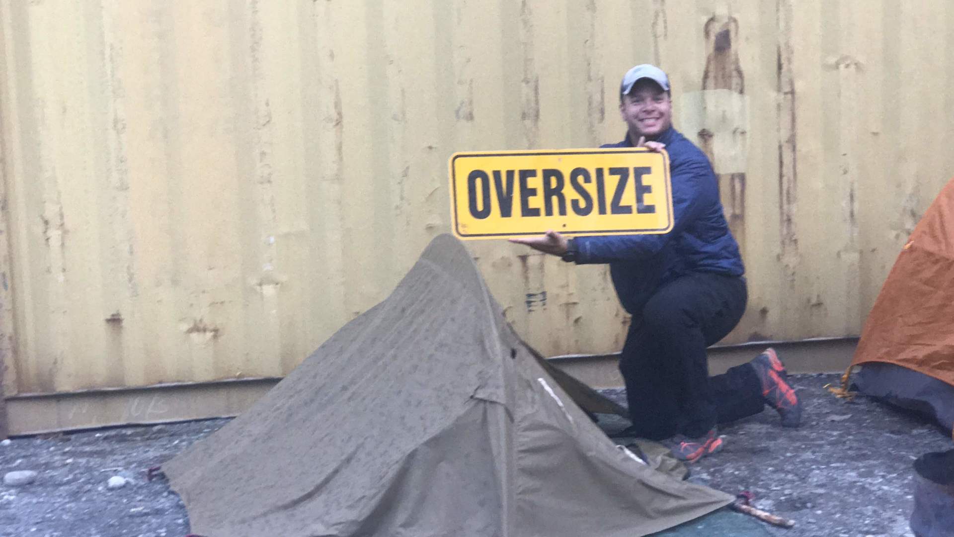 Person holding an oversized yellow sign beside a gray tent in front of a beige wall