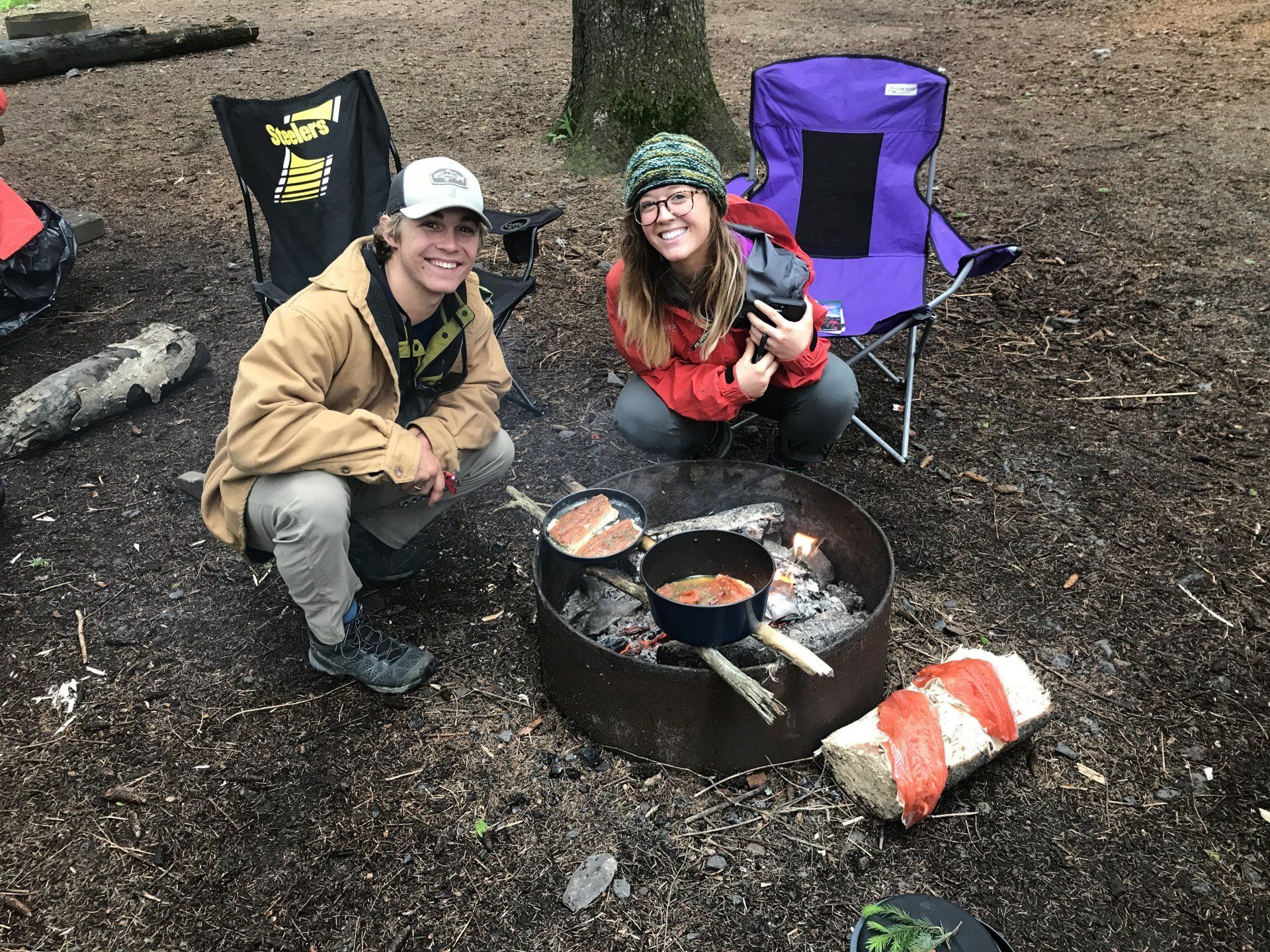 Two people sitting by a campfire with cooking pans and camping chairs in a wooded campsite.