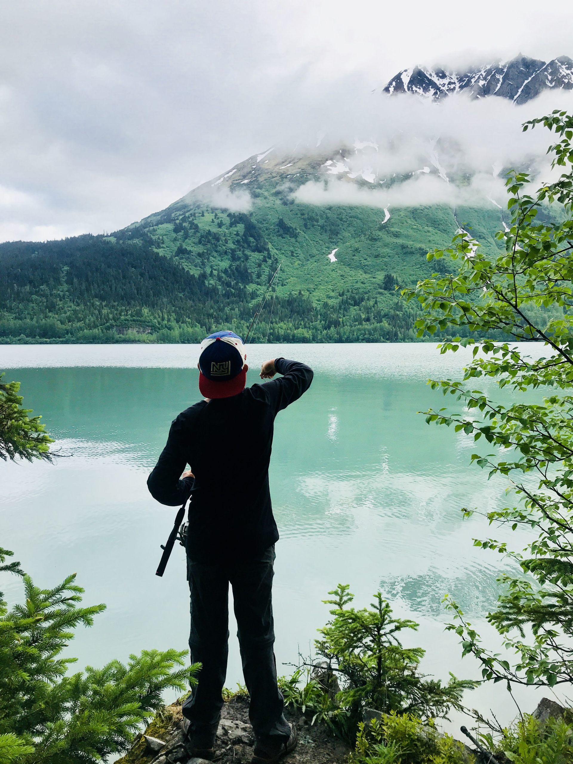Person in black stands by a turquoise lake, pointing toward misty green mountains.