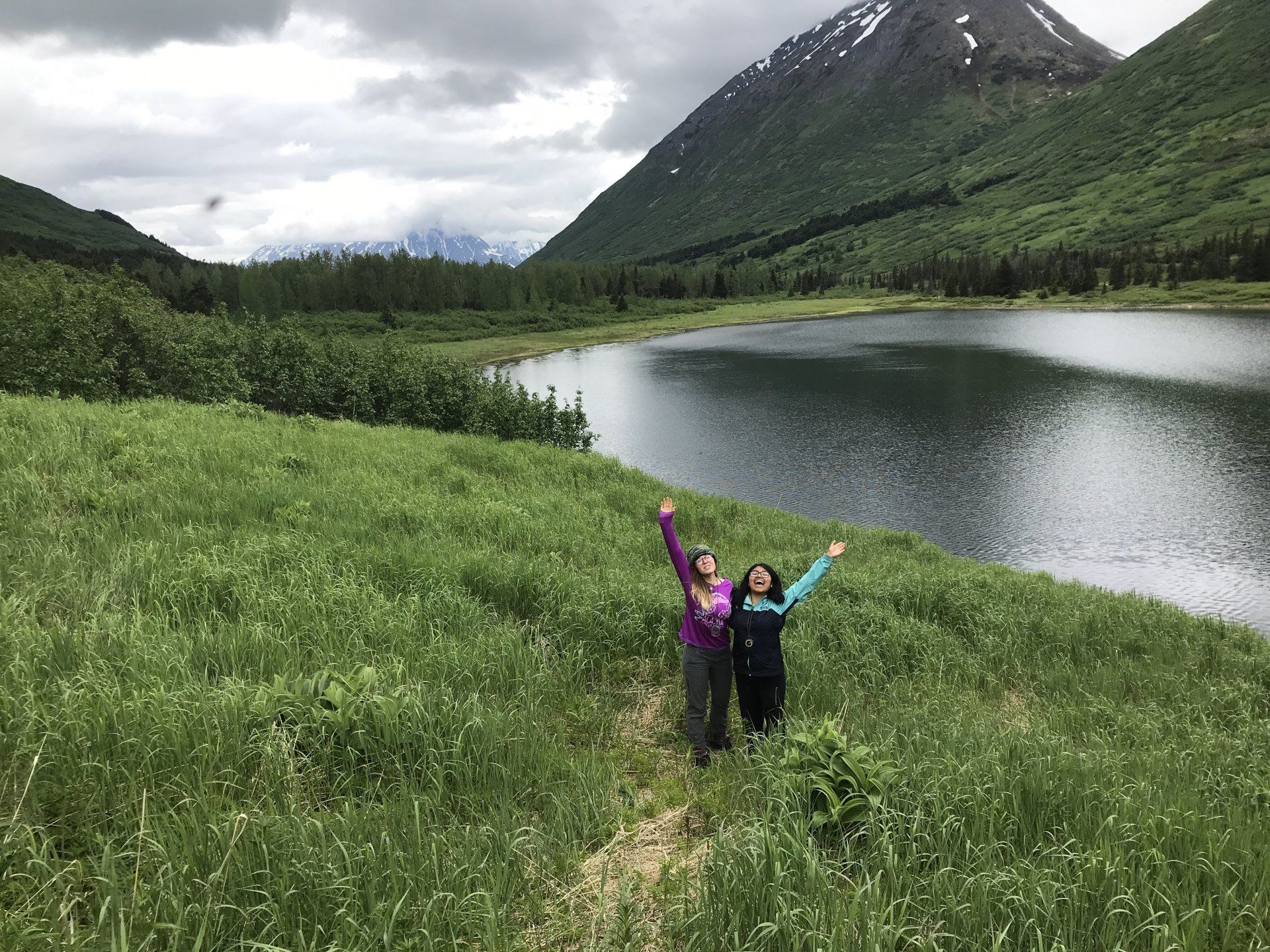 Two hikers with raised arms on a grassy trail beside a lake and mountain under cloudy skies
