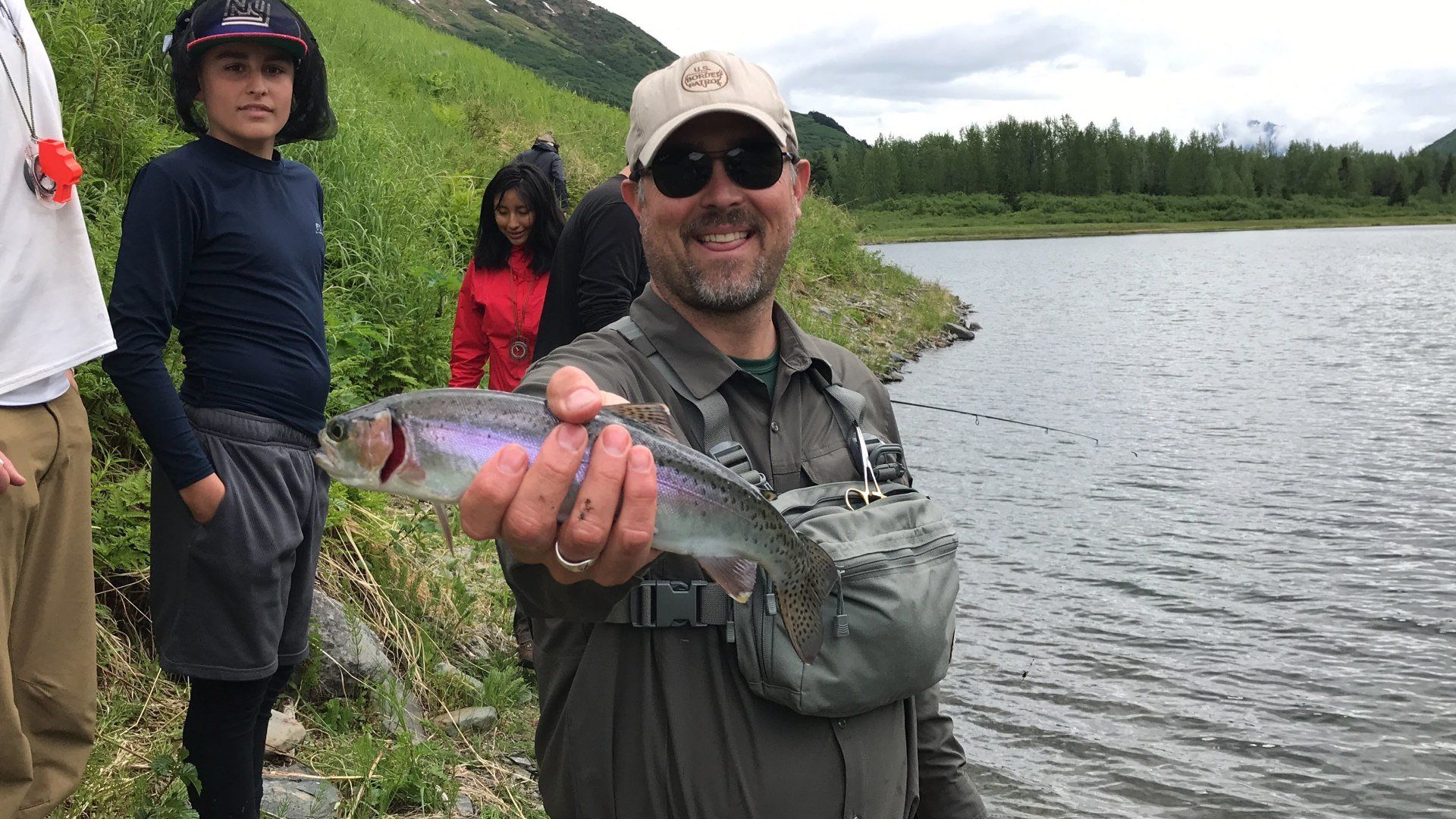 Smiling angler holding a fish by a lakeshore, with others standing nearby and water in the background