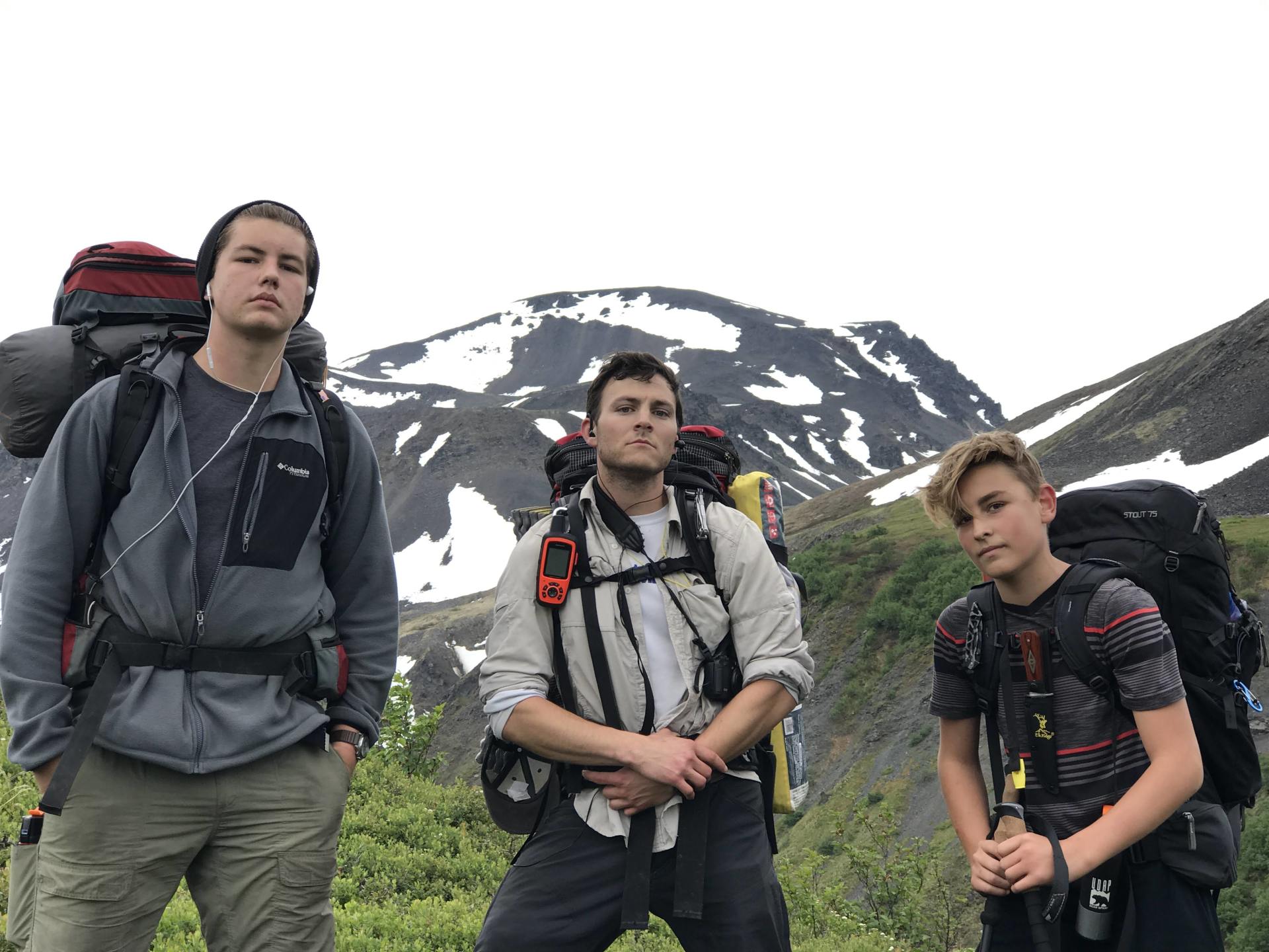Three hikers with backpacks standing on a rocky mountain trail with snow-covered peaks behind them