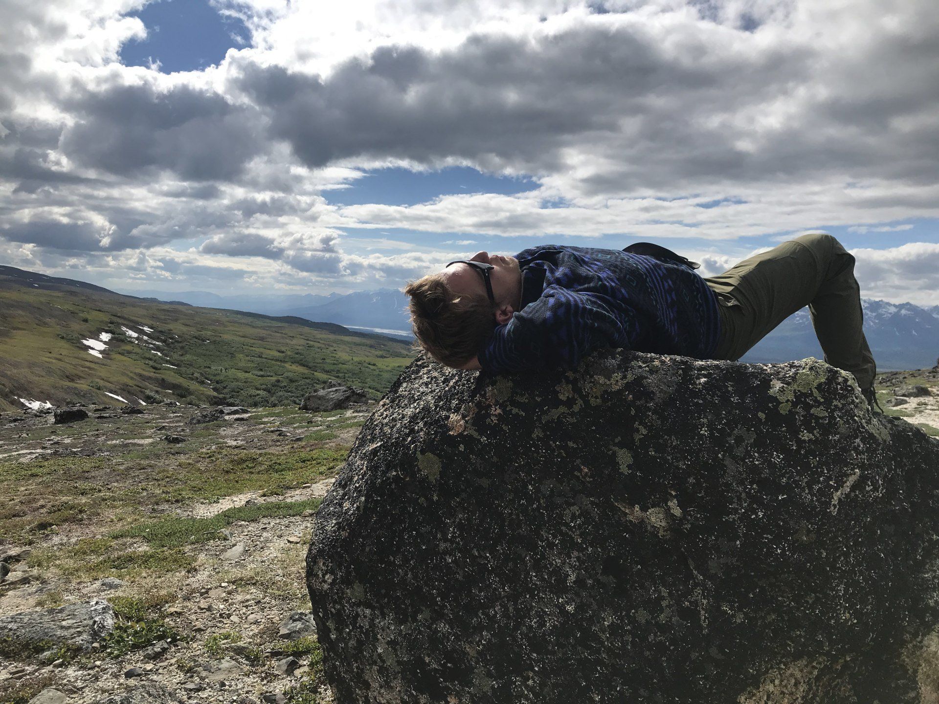 Person lying on a large rock in a windy mountain landscape under cloudy skies.