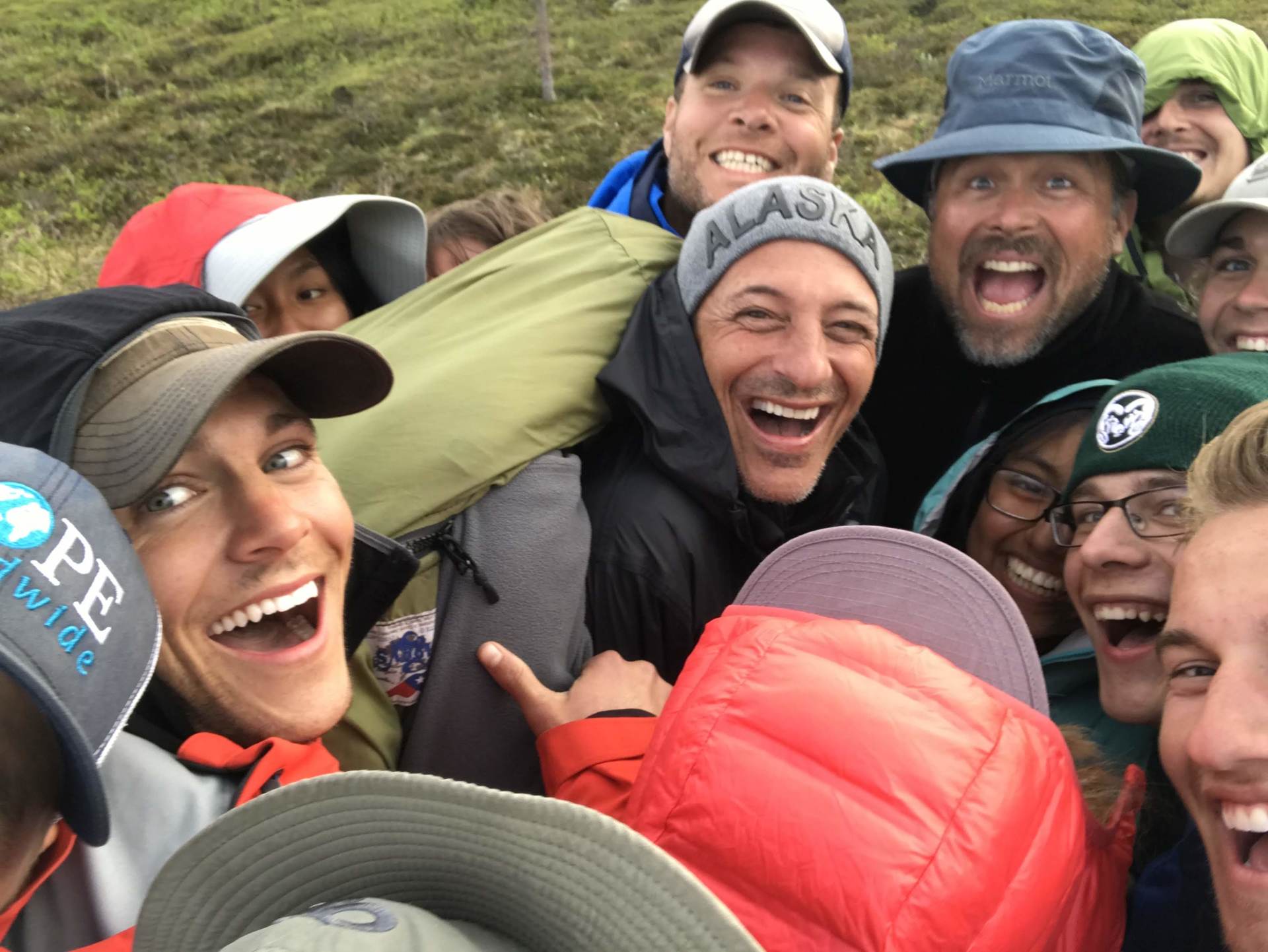 Smiling hikers huddled together outdoors, wearing colorful jackets and caps on a grassy hillside.