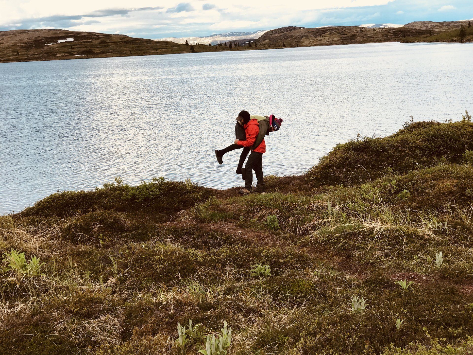 Two people in red jackets hiking beside a lake with grassy hills and cloudy sky