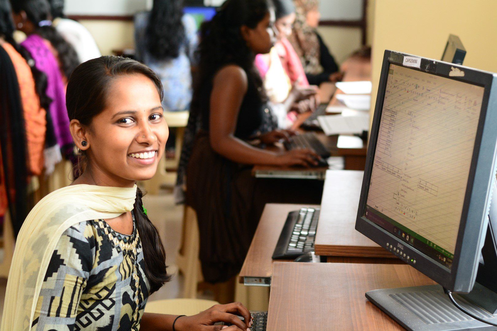 Woman smiling at a computer in a classroom, with several people working at laptops in the background.