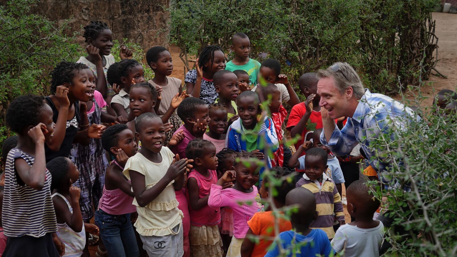 Man kneels among smiling children outdoors, reaching out in a colorful community scene.