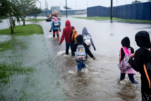 People wading through floodwater on a flooded sidewalk near blue buildings