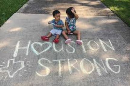 Two children sitting on a sidewalk beside chalk writing that says “Houston Strong” and a Texas outline
