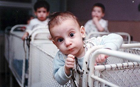 Baby leaning over a crib rail, looking at the camera; two children blurred in the background.