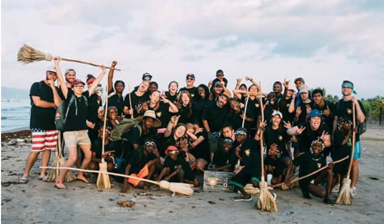 Large group posing on a sandy beach, many holding brooms and wearing dark shirts at sunset