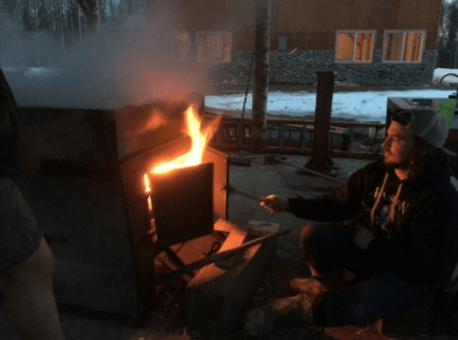Person sitting by a small outdoor fire at dusk, with snow on the ground and a cabin in the background