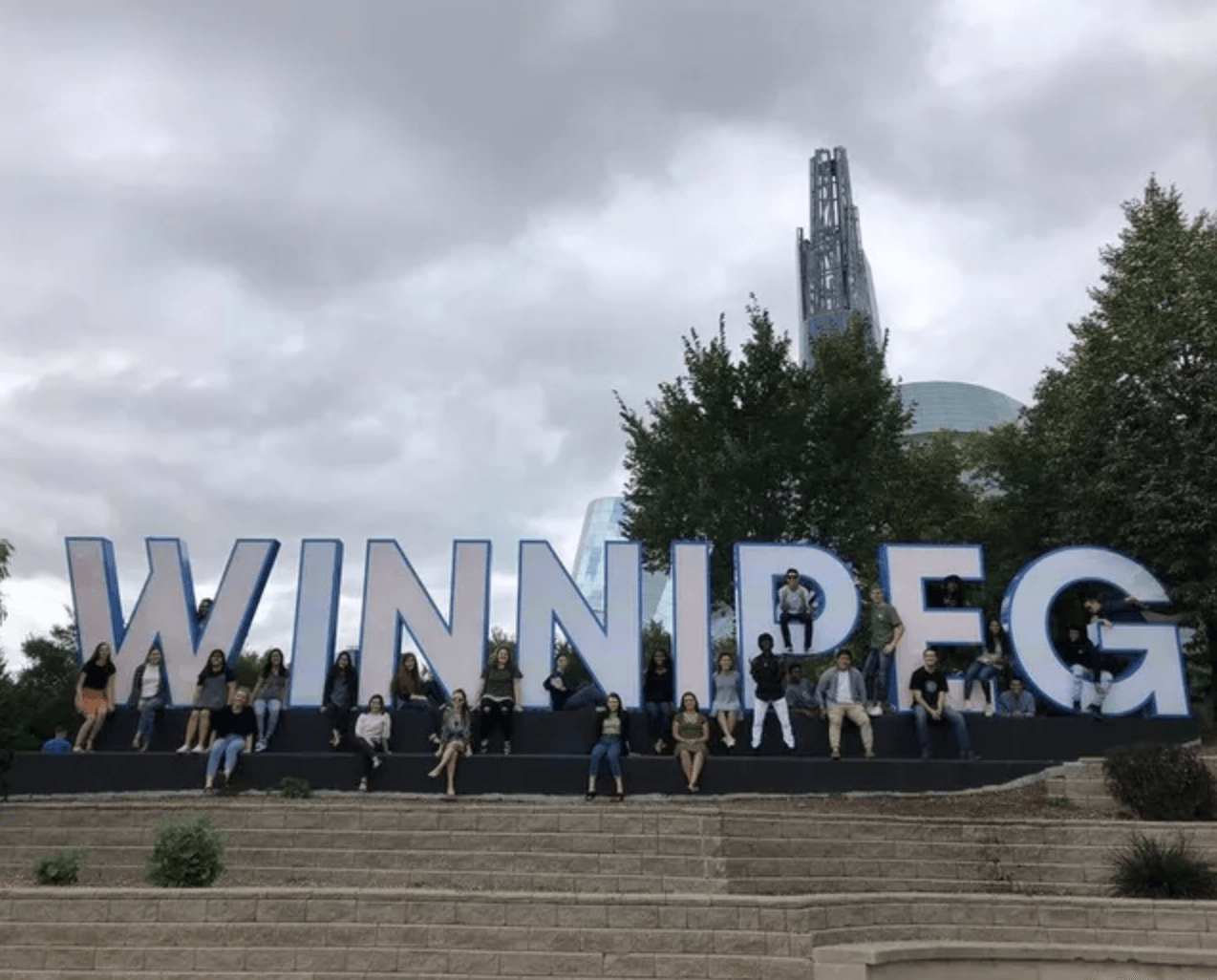Large “WINNIPEG” sign with people posing on steps under a cloudy sky, city tower in background
